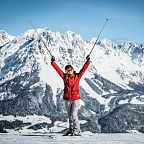 Skifahrer in roter Jacke hebt lächelnd die Arme vor verschneitem Bergpanorama mit blauem Himmel. Perfekte Winterlandschaft für Wintersport.