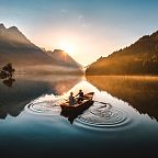 Two people rowing a boat on a calm lake at sunrise, surrounded by mountains, with the sun rising between peaks creating a serene and reflective atmosphere.