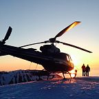 Silhouette of a helicopter and two people on a snowy peak at sunset in Wilder Kaiser, with orange and pink sky highlighting the tranquil winter scene.