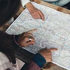 Personen schauen auf eine Landkarte auf einem Tisch, während sie mit Fingern Richtungen zeigen. Eine entspannte Outdoor-Szene in einem natürlichen Setting.