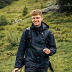 Person in dunkler Outdoor-Kleidung wandert mit einer Kamera in der Hand durch eine grüne, leicht hügelige Landschaft.