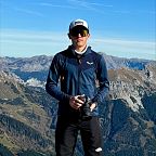 Ein Mann steht auf einem Berg mit Kamera in der Hand, umgeben von einer beeindruckenden Alpenlandschaft mit blauen Himmel und schneebedeckten Gipfeln.