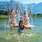 Three people, including a child, splash in a clear mountain lake, set against the stunning backdrop of the Wilder Kaiser mountains under a bright blue sky.