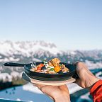 Eine Pfanne mit Kaiserschmarrn, serviert vor einer verschneiten Berglandschaft bei klarem Himmel. Der winterliche Hintergrund bietet eine beeindruckende Aussicht.