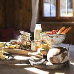 A rustic wooden table set with traditional Tyrolean foods: fresh bread, cheese, selection of vegetables, and a bottle of milk, inside a warm, sunlit alpine hut.