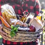 A person holding a wicker basket filled with fresh groceries including pasta, a carrot, eggs, cheese, a tomato, and a basil plant.