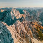 Luftaufnahme eines beeindruckenden Bergmassivs bei Sonnenaufgang mit schroffen Felsen und Tälern, die in sanftem Licht getaucht sind.