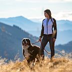 Frau in sportlicher Kleidung steht mit einem Hund auf einem Berg, umgeben von malerischen Bergen und blauem Himmel im Hintergrund an einem sonnigen Tag.