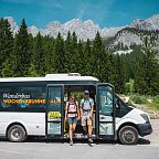 Hiking bus with people stepping out in front of the majestic Wilder Kaiser mountains, surrounded by lush greenery and rugged peaks under a clear blue sky.