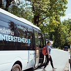 A woman with a backpack boards a hiking bus to Hintersteiner See in the Wilder Kaiser region. The bus is parked on a road surrounded by green trees and a blue sky.