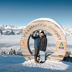 Two people stand by a wooden circle in a snowy landscape at Wilder Kaiser, Austria, with mountain views and clear blue skies.