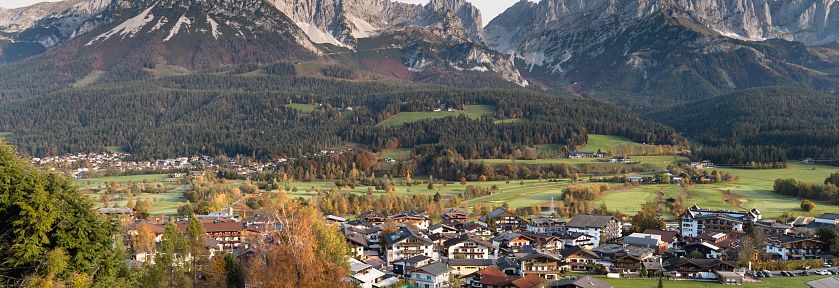 Blick auf ein Tiroler Dorf im Herbst, umgeben von üppigen Wäldern und dem beeindruckenden Bergmassiv des Wilden Kaisers im Hintergrund.