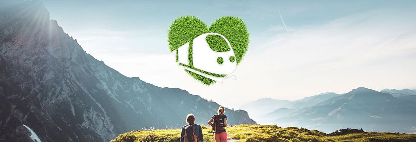 Zwei Wanderer auf einem Bergpfad, umgeben von üppiger grüner Natur und einem Bergpanorama. Ein Zug in Form eines Herzlogos symbolisiert umweltfreundliches Reisen.