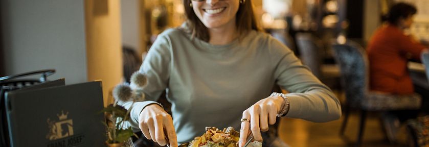 Frau in einem Restaurant, die lächelnd ein Gericht mit Besteck isst. Im Hintergrund ist eine gemütliche, warme Atmosphäre zu erkennen.