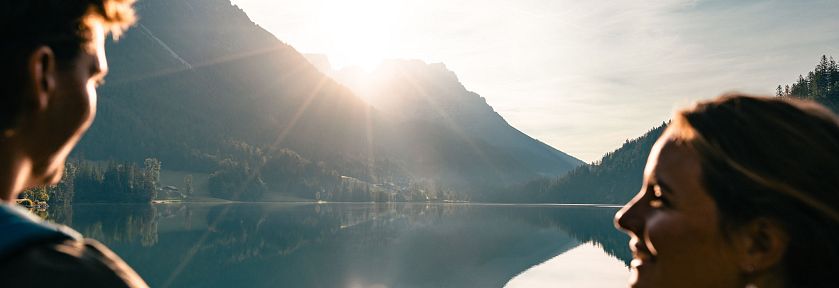 Ein Paar steht am Ufer eines ruhigen Sees, der die umliegenden Berge und den klaren Himmel reflektiert, während die Sonne hinter den Gipfeln hervorkommt.