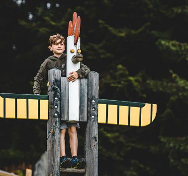 Junge steht hinter Hahn ähnlicher Holzskulptur und hält sich an dieser fest