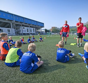 Zwei Trainer in rotem Trikot sprechen zu im Halbkreis sitzenden Kindern