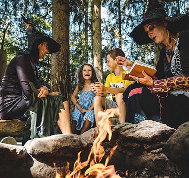 Als Hexen gekleidete Frauen sitzen mit Kindern um Lagerfeuer und lesen aus einem Buch vor