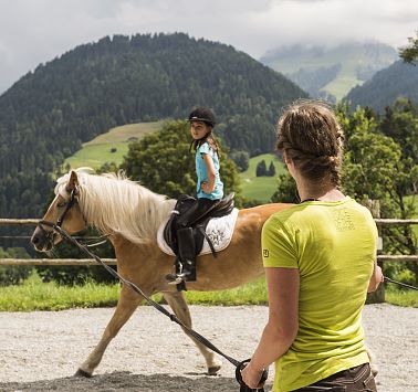 Mädchen auf hellbraunem Pferd wird von Reitlehrerin an der Pferdeleine geführt