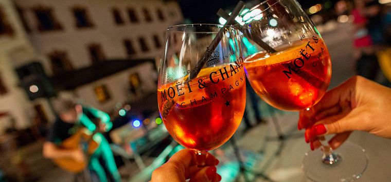 Zwei Hände stoßen mit Aperol an im Hintergrund Musikband